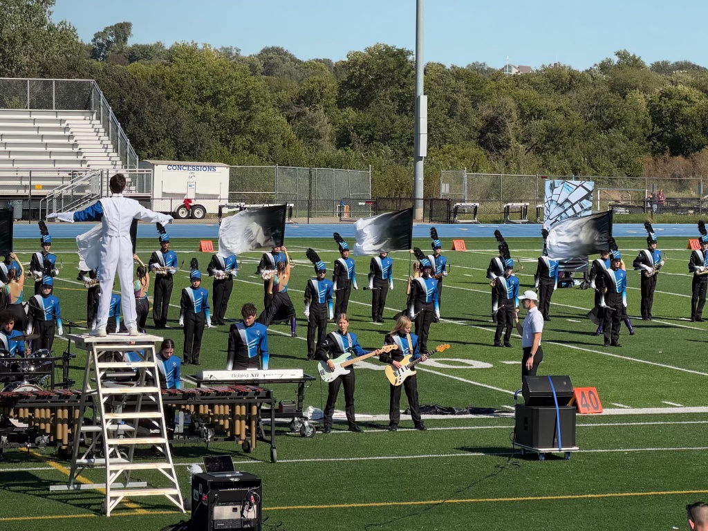 A marching band performance on a football field, featuring a conductor standing on a ladder, musicians playing instruments, and color guard members with flags in motion.