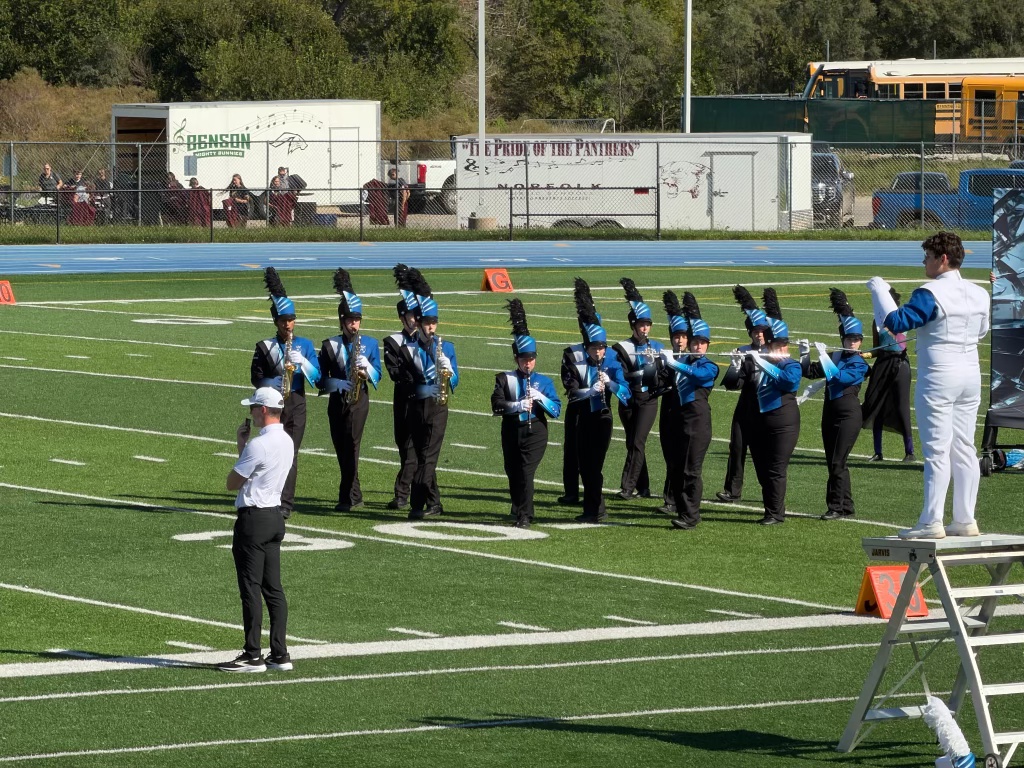 A marching band performs on a football field, with members dressed in blue and black uniforms, some wearing tall feathered hats. A conductor stands on a platform, directing the group.