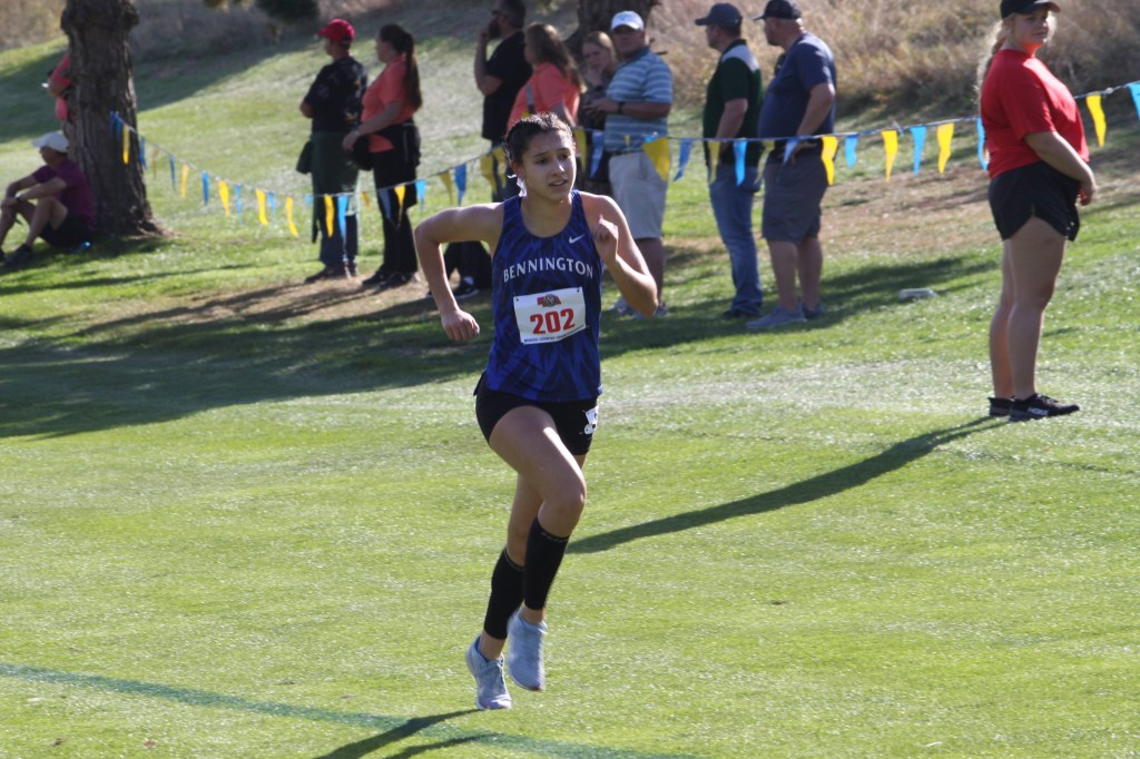 A female cross-country runner wearing a blue jersey with 'BENNINGTON' and a number '202' accelerates on a green grass field, with spectators in the background.