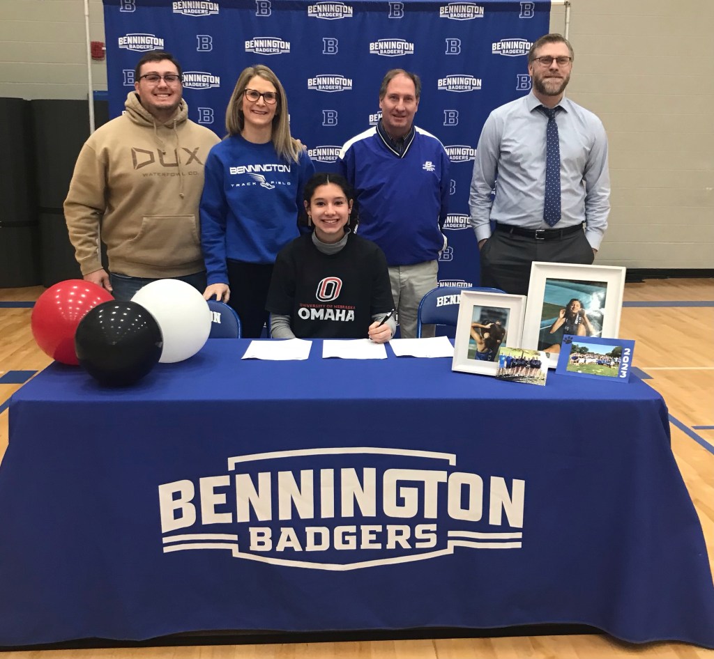 A group of individuals celebrating a student-athlete's signing at a table decorated with balloons, featuring 'Bennington Badgers' branding. The student is signing a letter of intent while surrounded by family and school staff.