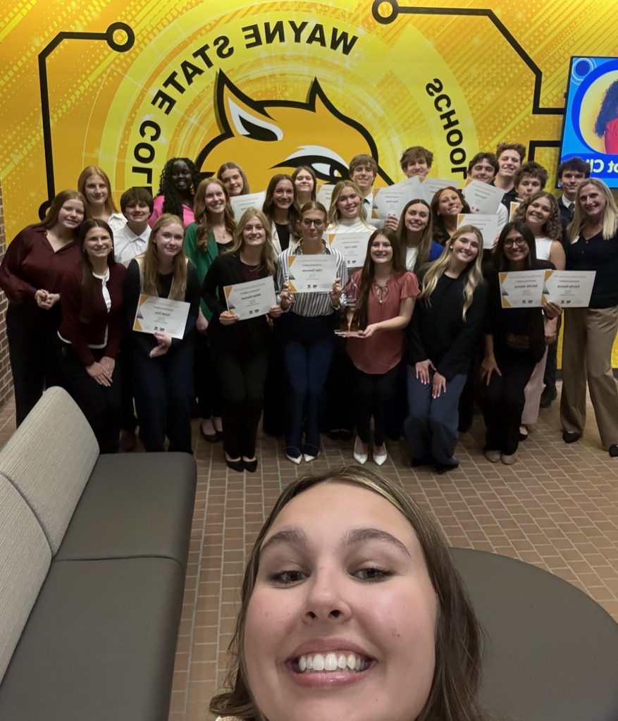 A smiling individual takes a selfie while a group of students stands behind, holding certificates and a trophy, in front of a colorful Wayne State College backdrop.