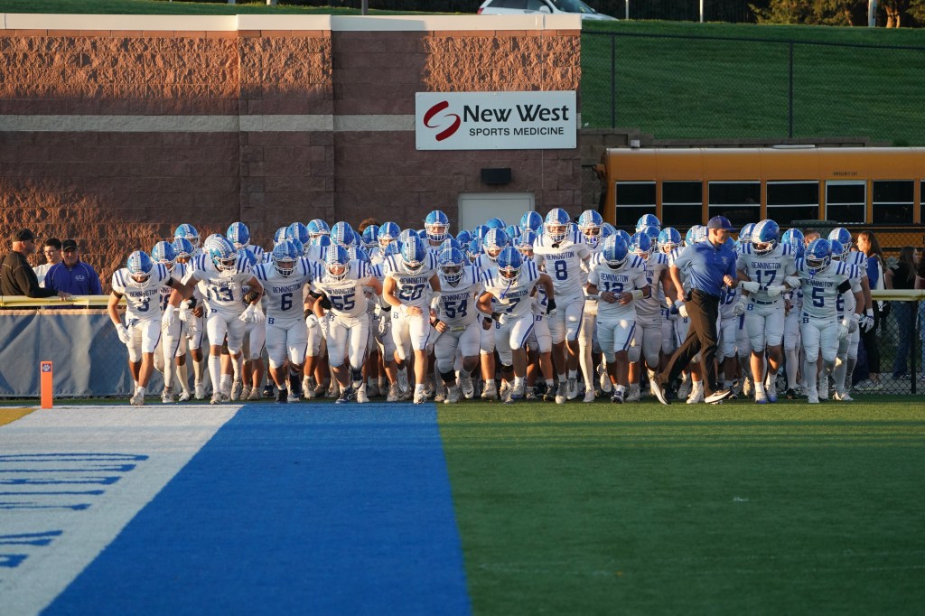 A football team in white and blue uniforms running onto the field, with a coach leading them. A school bus is visible in the background.