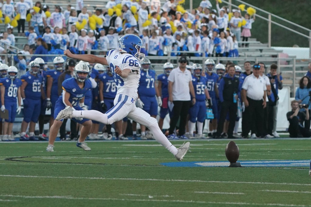 A high school football player executing a kick on the field, with teammates and fans in the background.