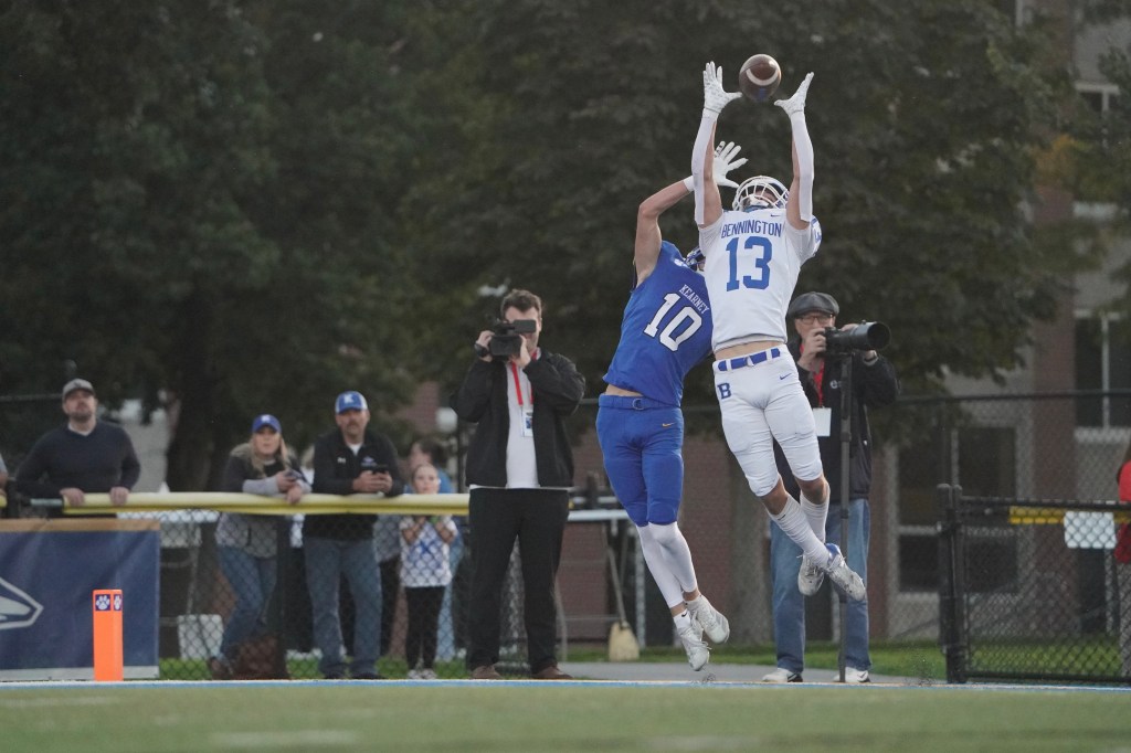 A football player in a blue uniform leaps high to catch the ball while another player in a white uniform attempts to defend against the catch. Spectators are visible in the background, watching the game.