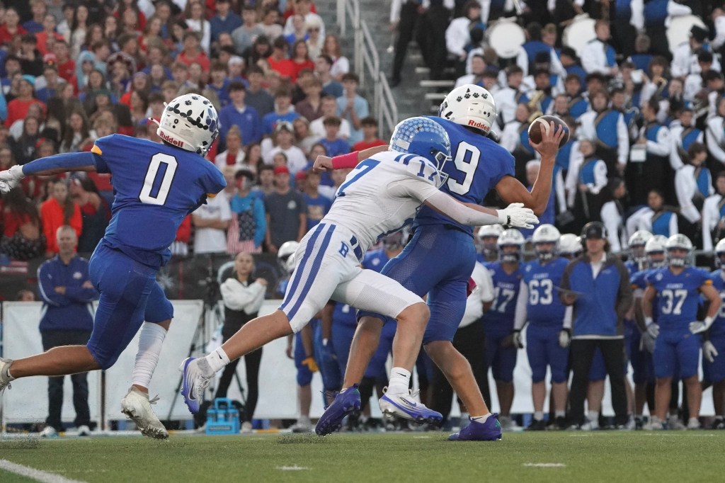 A football player in a blue uniform with the number 9 throws the ball while being pressured by a defender in a white uniform, number 17, who is attempting a tackle. Another player in blue with the number 0 is in the background. Spectators are visible in the stands.