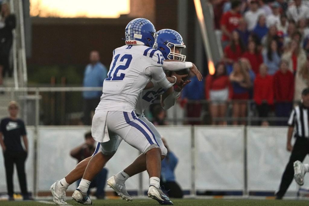 A football player in a blue helmet and white uniform hands the ball off to another player wearing a similar uniform during a game.