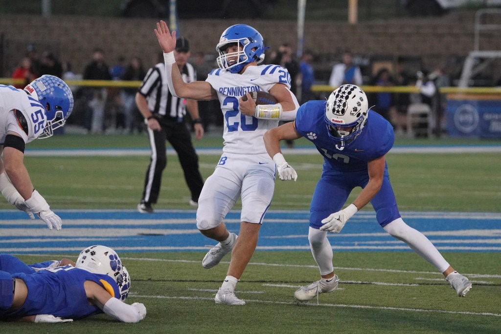 A football player in a white uniform attempts to evade a tackle by an opposing player in a blue uniform, with another player on the ground in a blue uniform during a game.