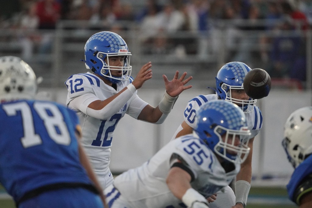 A high school football quarterback in a white uniform with blue accents, wearing number 12, throws a pass while players from both teams line up for a play.