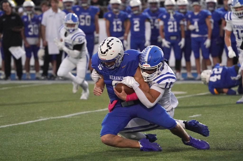 A football player in a blue jersey is being tackled by an opposing player in white and blue, on a field with other players in the background.