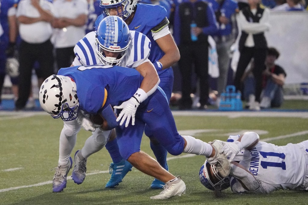 A football player in a blue uniform is being tackled by two opponents, one in a white and blue striped helmet and the other on the ground. The scene captures the intensity of the game, with spectators blurred in the background.