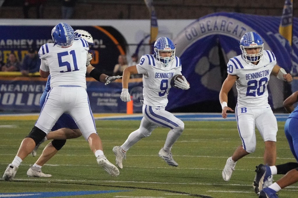 Three football players in white uniforms with blue stripes and 'Bennington' written on them, during a game on a turf field. Player number 9 is running with the ball while players 51 and 20 are providing support.