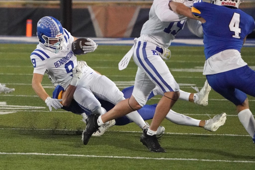 A football player in a white uniform with blue stripes is being tackled by an opponent while holding the ball during a game.