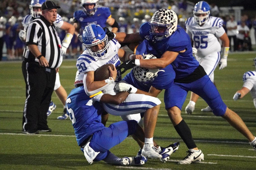 A football player in a blue uniform is being tackled by two opponents during a game, with a referee observing in the background.