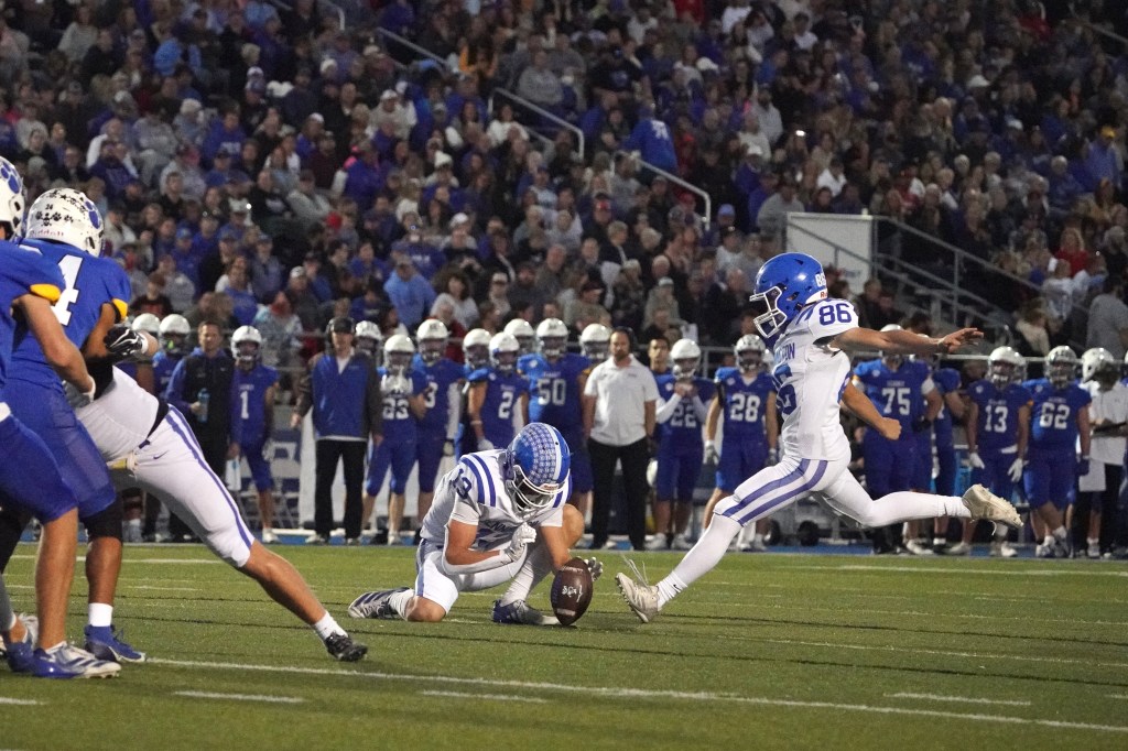 A moment during a football game featuring a player preparing for a kick while another player holds the ball on the field, surrounded by teammates and spectators in the background.
