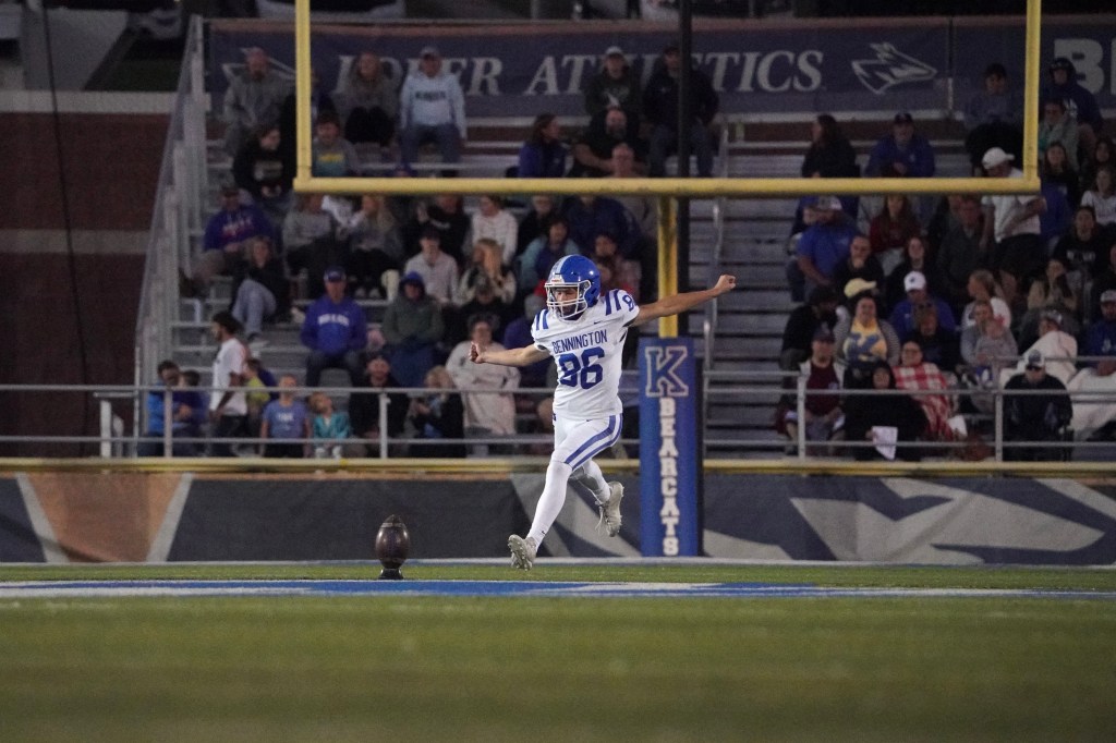 A football player in a white jersey with blue accents preparing to kick a football during a night game, with a crowd in the stands behind him.