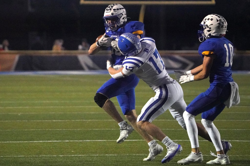 A football player in a blue uniform is being tackled by an opponent in a white and blue uniform on a grassy field during a night game.