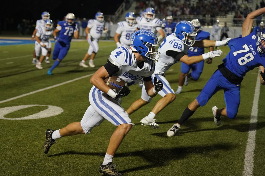 A high school football player wearing a white and blue uniform runs with the ball on a grassy field, while players from the opposing team in blue attempt to tackle him.