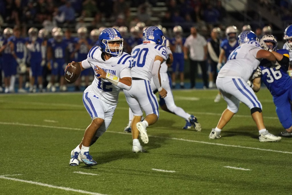 A high school football player in a white uniform with blue accents, wearing the number 12, prepares to throw the ball while evading defenders during a night game.