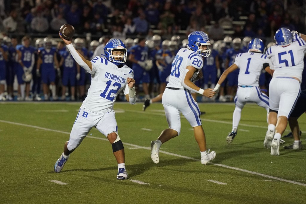 A football player wearing a white jersey with 'BENNINGTON' and number 12 is throwing a pass while teammates run in the background during a night game.