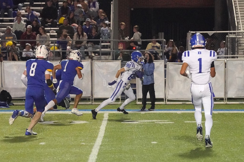 High school football players on the field during a night game, with one player in blue running toward the end zone while others pursue him.