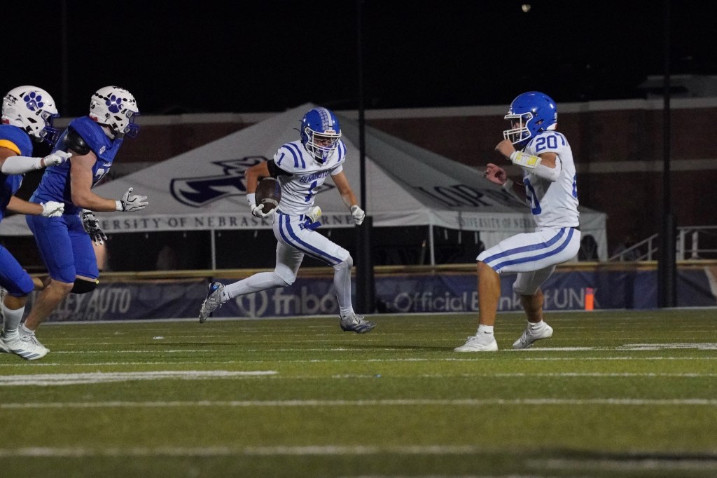 A football player in a white jersey with blue stripes carries the ball while two defenders in blue jerseys are attempting to tackle him during a night game.