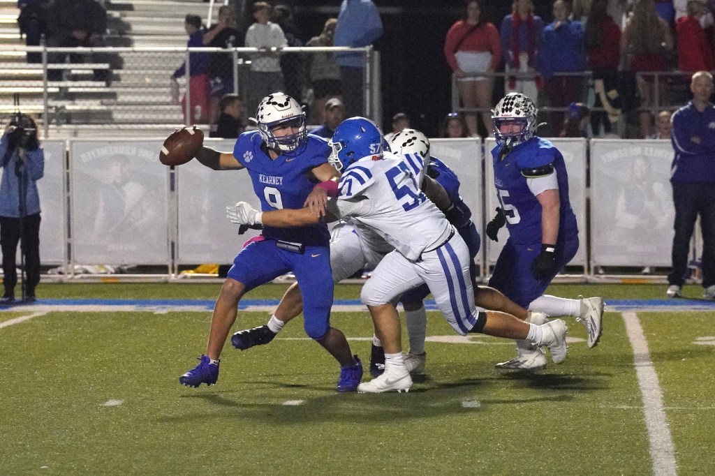 A football player in blue is attempting to throw the ball while being chased by a defender in white. The scene takes place on a football field, with spectators in the background.