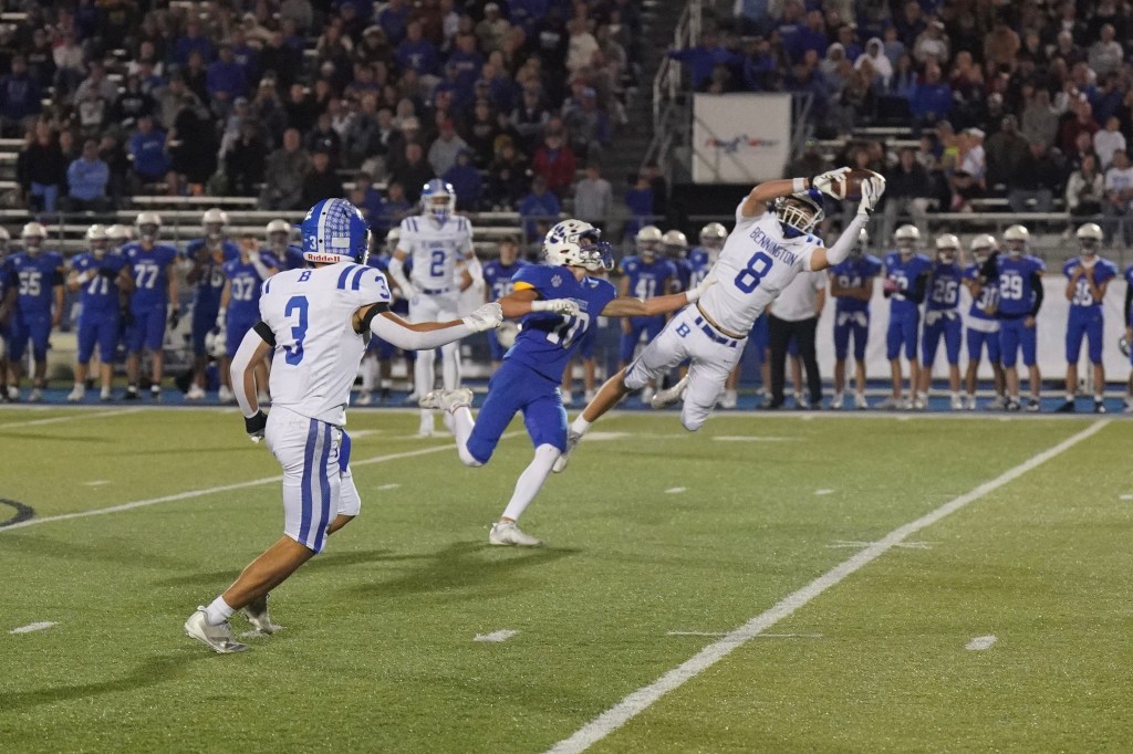 A football player in white and blue uniform leaps to catch a pass during a game, with a blue team player attempting to defend. Spectators watch in the background.