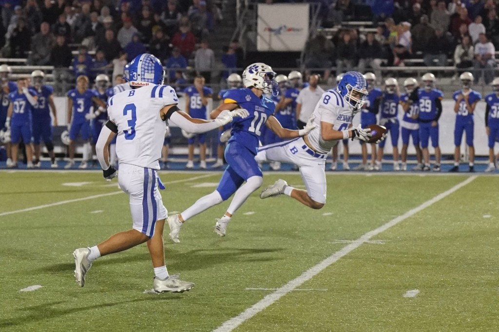 A football player wearing a white jersey with blue accents catches the ball while another player in a blue jersey attempts to tackle him on the field. A crowd is visible in the background.