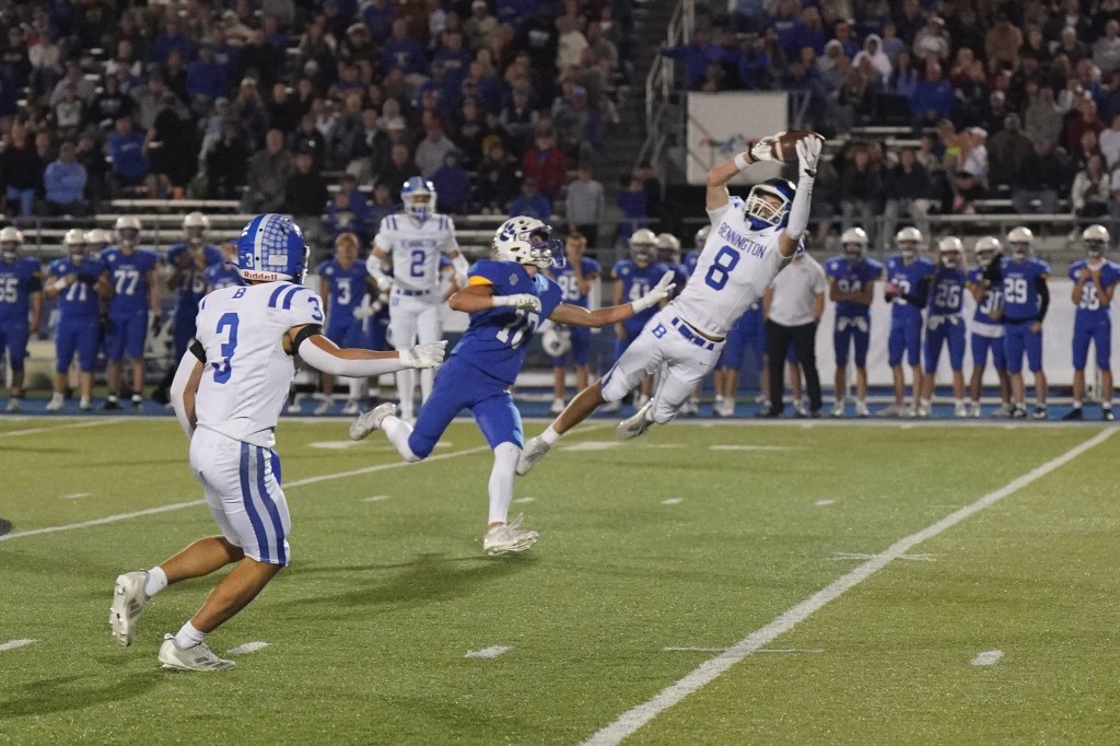 A high school football player in a white uniform with blue accents makes an impressive catch while jumping in mid-air. Another player in a blue uniform is attempting to defend but is unable to reach the ball. The background shows a cheering crowd and other players on the sidelines.