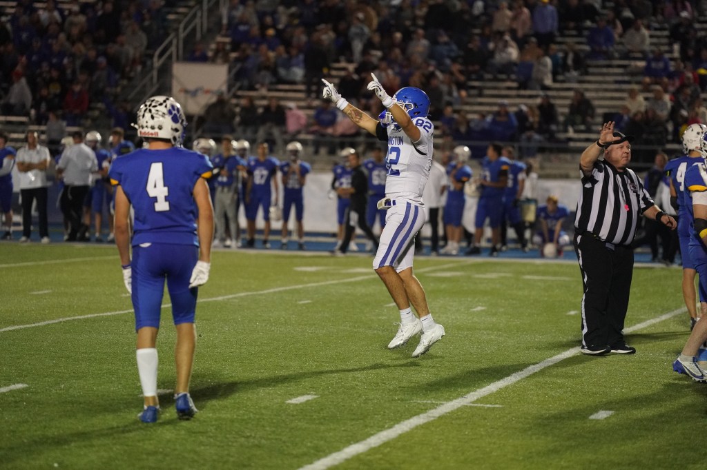 A football player with the number 82 celebrates in mid-air during a game, while another player with the number 4 stands nearby. A referee signals on the right side, and the crowd is visible in the background.