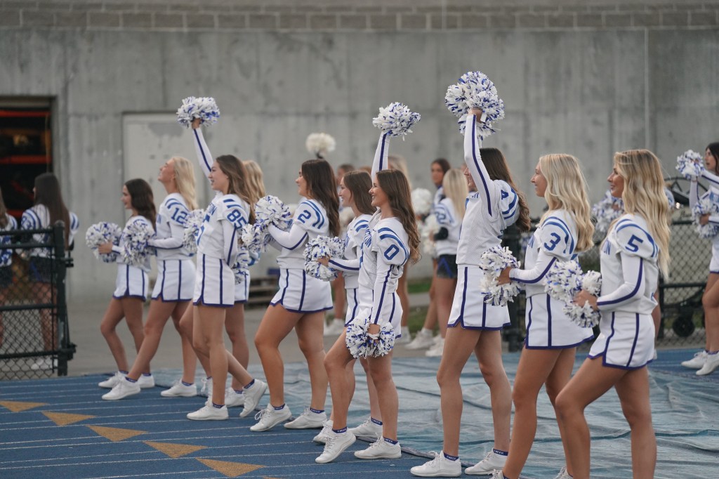 A group of cheerleaders in matching uniforms with blue and white patterns, holding pom-poms and performing a routine on a sports field.