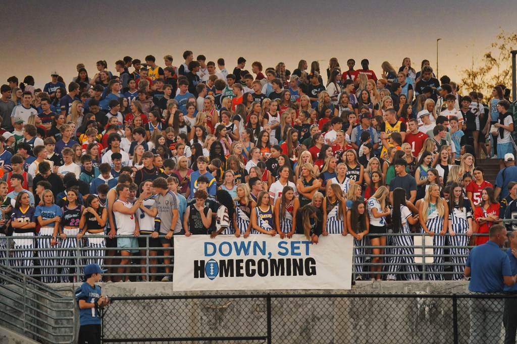 A large crowd of high school students gathered at a sports event, wearing various team jerseys and school colors, with a banner reading 'DOWN, SET, HOMECOMING' prominently displayed.