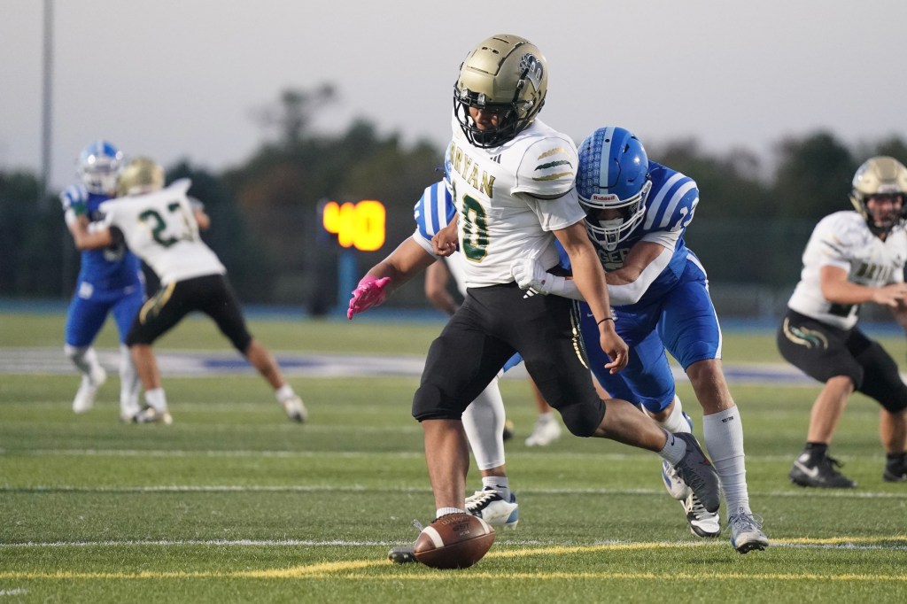 A football player in a white jersey with green accents is dropping the ball while being tackled from behind by an opponent in a blue jersey. The background shows other players on the field and a scoreboard.