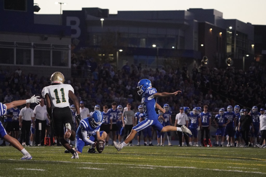 A high school football game in progress at dusk, with a player in blue uniform (#86) preparing to kick a field goal while another player in green (#11) runs towards him. A crowd is visible in the background.