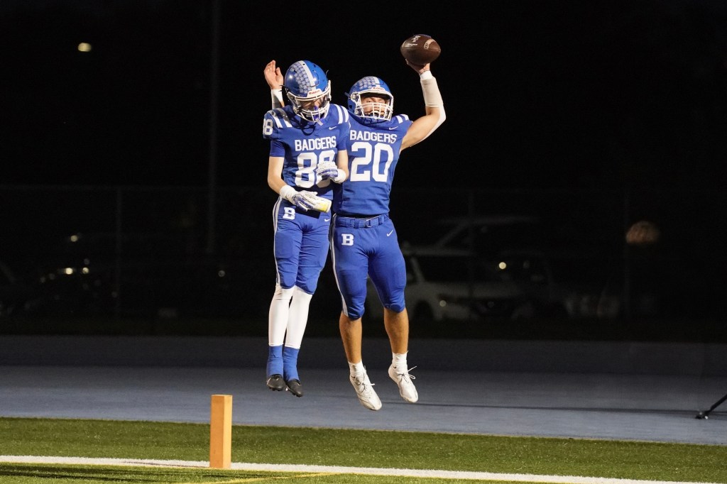 Two football players in blue uniforms celebrate on the field, one is jumping with a football raised in the air while the other is giving a high five.