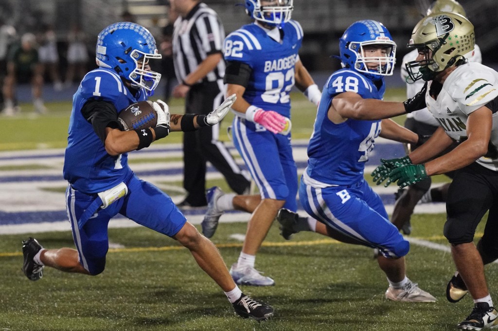 A high school football game scene with players in blue and white uniforms, one player running with the ball while being approached by a defender in green and white. Teammates are visible in the background.