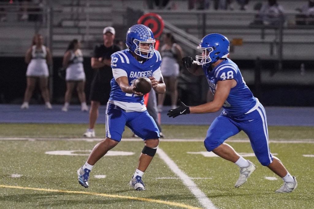 Two football players in blue uniforms executing a play on a field under stadium lights, with a referee and cheerleaders in the background.