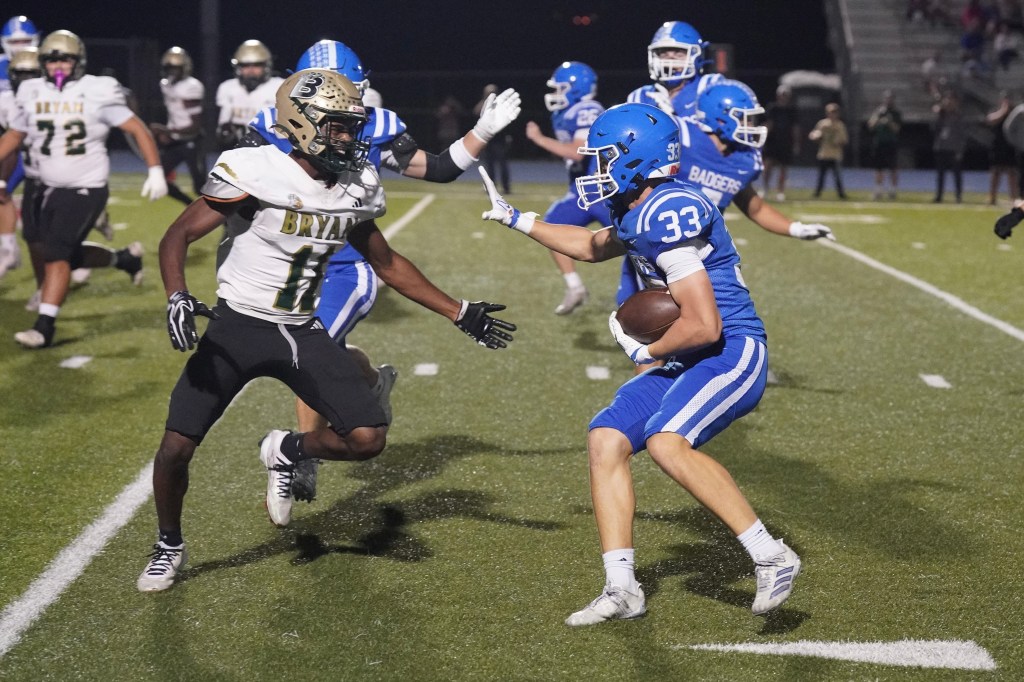 A football player in a blue uniform evading a tackle from an opponent in a white and gold uniform on a grass football field during a night game.