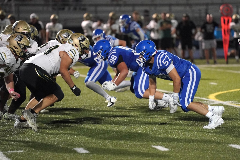 Two football teams lined up on the field, one in blue jerseys and the other in white and gold. Players are in a ready stance, preparing for the snap.