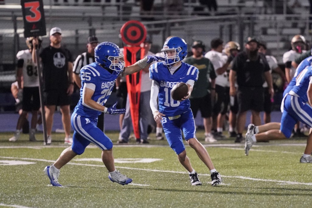 A high school football game with players wearing blue uniforms. One player, number 25, is preparing to receive a handoff from the quarterback, number 10, who is in the process of passing the ball.