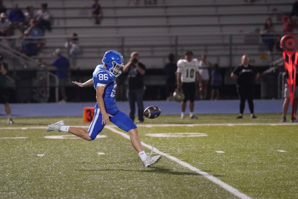 A high school football player in a blue uniform, number 86, executing a kick on a field during a night game, with spectators in the background.