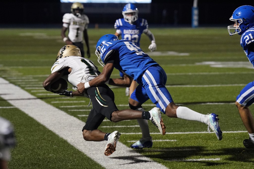 A football player in black and gold is being tackled by a player in blue during a night game on a football field.