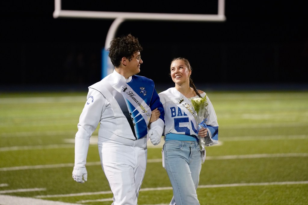 A young man wearing a band uniform and a sash walks arm in arm with a young woman dressed in a jersey, both smiling during a homecoming event on a football field.