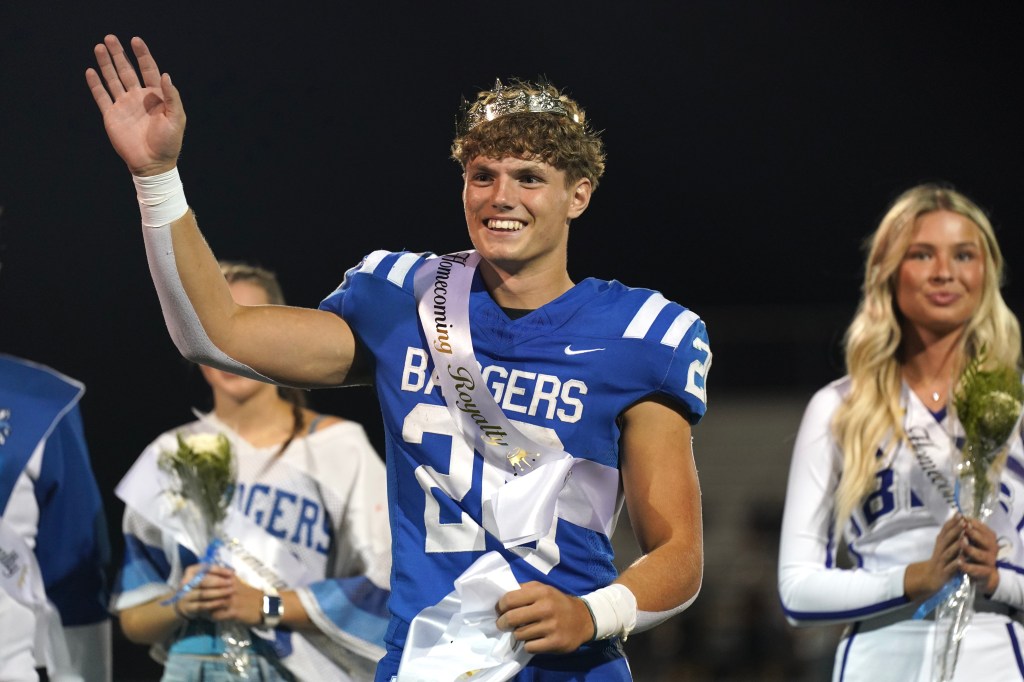 A young male football player wearing a blue jersey with 'Badgers' printed on it and a homecoming sash, waving to the crowd during a celebration, with a female student holding flowers next to him.