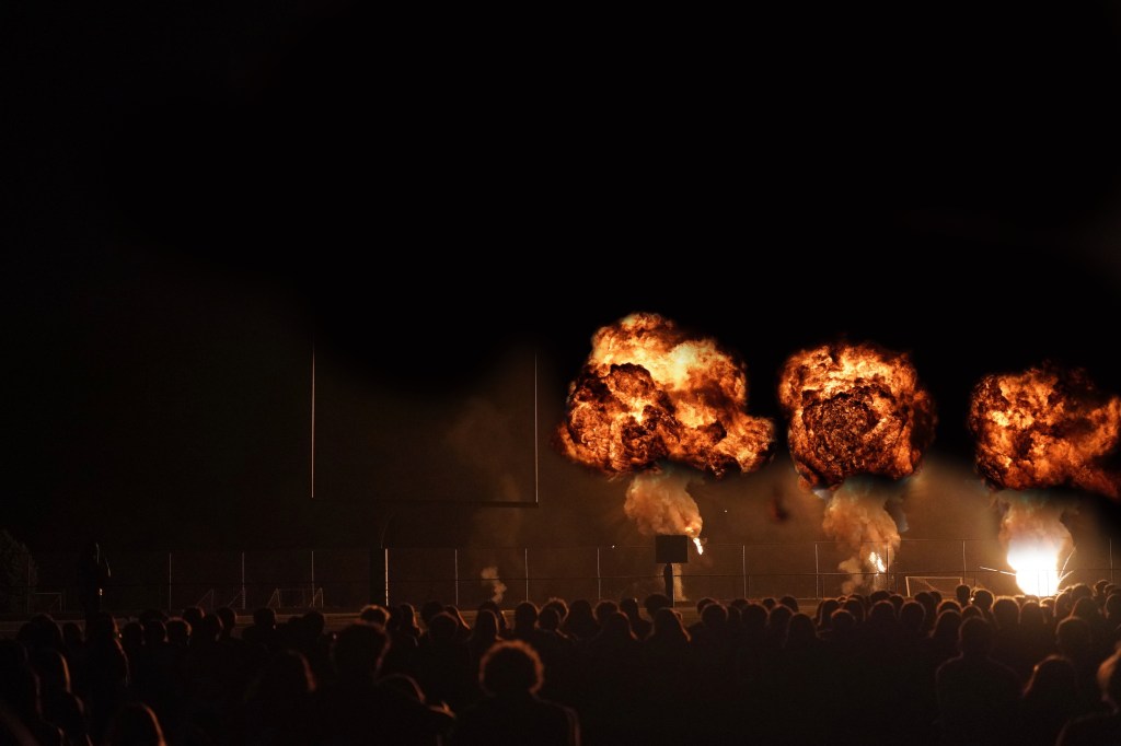 A crowd watching a nighttime display of explosions with flames and smoke in the background, featuring a football goalpost.