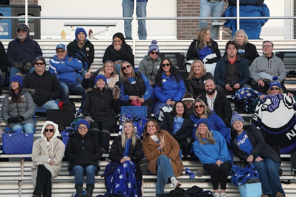 A group of fans cheerfully seated in a stadium, dressed in blue and black attire, some with blankets and hats, enjoying a sporting event.