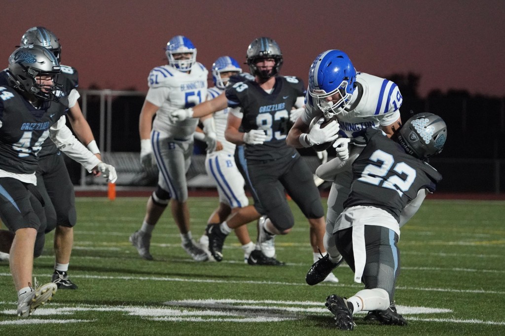 A high school football game scene with players in black and blue uniforms in an action-packed moment, as one player attempts to tackle another on an artificial turf field.