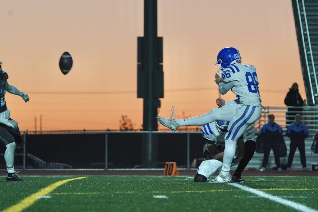 A football player in a white uniform with blue accents is about to kick a football during a game. The scene is set against a sunset sky, with other players and a stadium in the background.