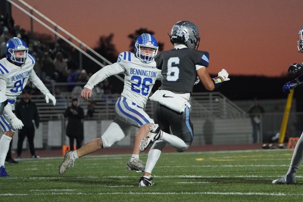 A high school football game scene at dusk, featuring a player in a black uniform with the number 6 running away from a defender in a blue and white uniform representing Bennington. Two other players can be seen in the background, and the atmosphere is charged with competition.