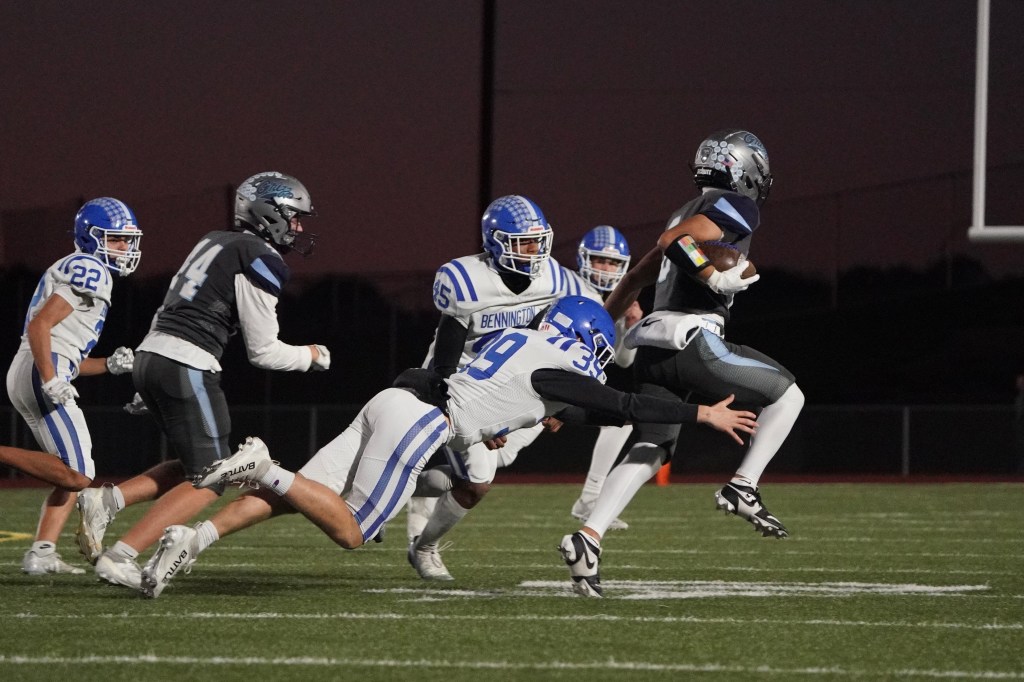 A high school football player in a gray uniform dodges a tackle from a defender in a white and blue uniform while several other players look on during a night game.